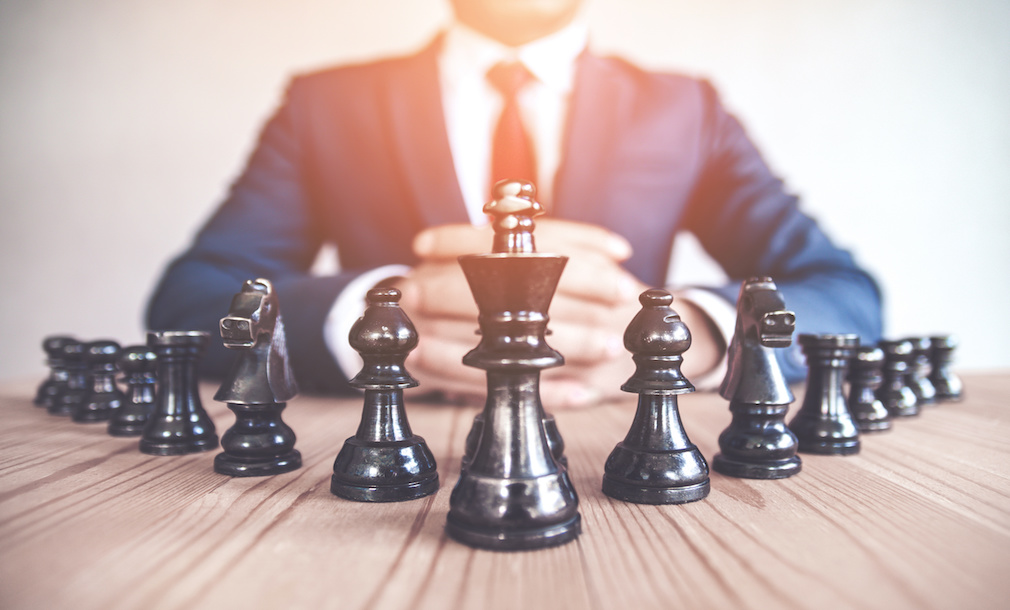 Retro style image of a businessman with clasped hands planning strategy with chess figures on an old wooden table.