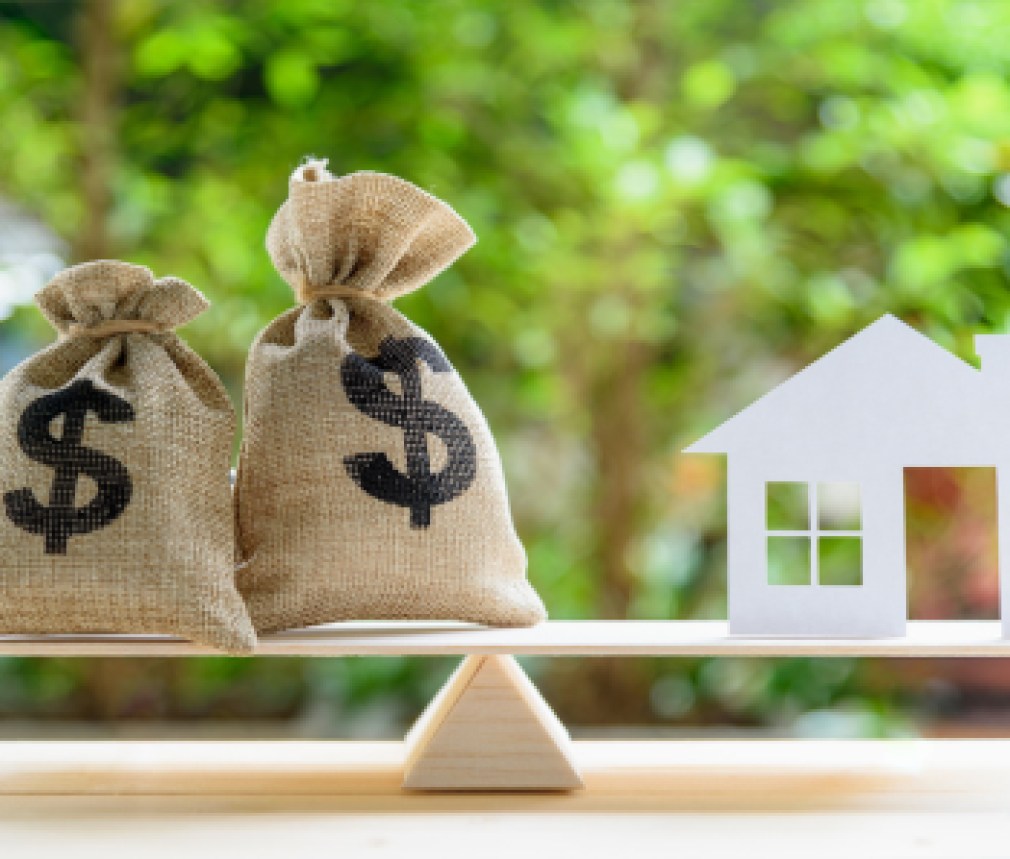 Home loan / reverse mortgage or transforming assets into cash concept : House paper model , US dollar hessian bags on a wood balance scale, depicts a homeowner or a borrower turns properties into cash